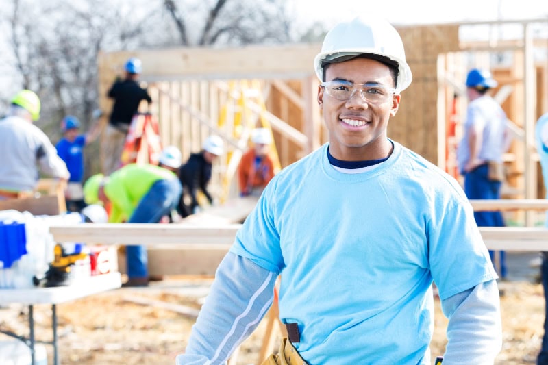 Schedule Your Annual Furnace Inspection Now - Handsome African American construction foreman or building contractor stands in front of home that is being built for charity. Volunteers are working on the home in the background. He hsi wearing a hard hat