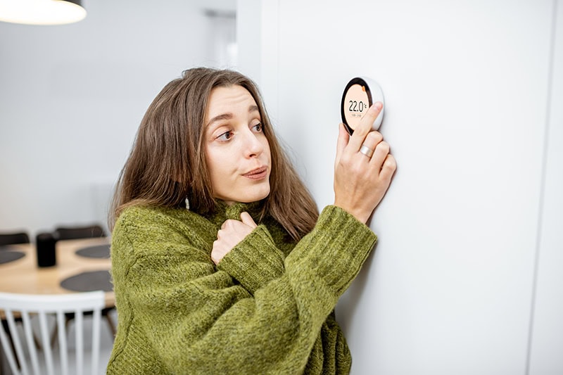 woman shivering in sweater as she adjusts her thermostat.